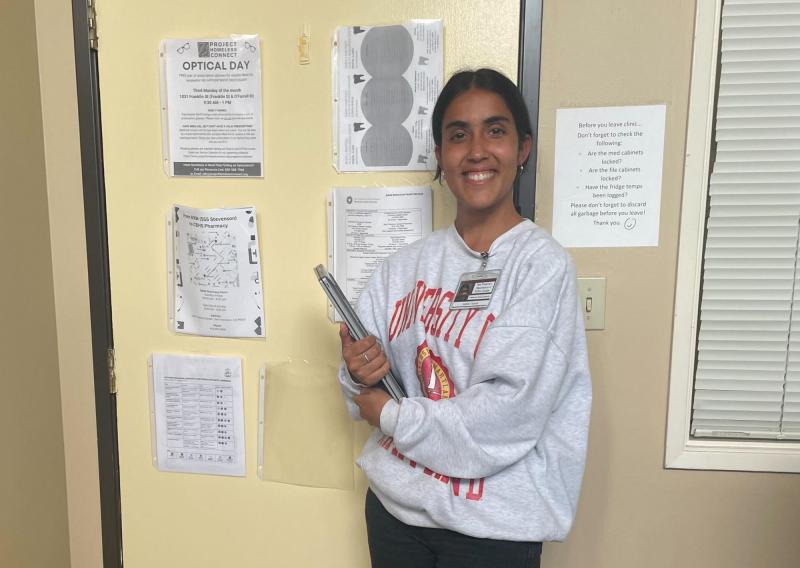 Anjalie is off-center of the frame, facing the camera and smiling. She is holding a resources binder. 