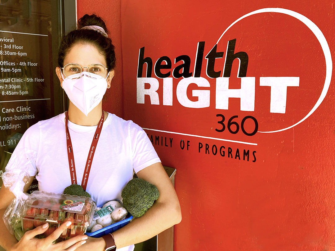 Mariana Carranza is seen standing in front of a HealthRIGHT 360 sign. She is holding assorted produce to give to patients.